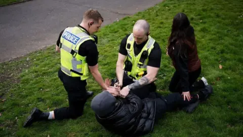 Two police officers pinning someone on a grass floor and handcuffing them. The person on the floor is wearing a black hoodie and outfit. A woman is next to him.