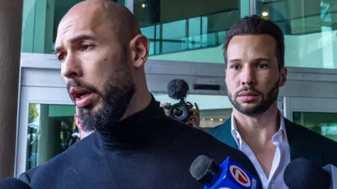 EPA Andrew Tate and his brother Tristan at Fort Lauderdale Airport in Florida with media pointing microphones at them for comment