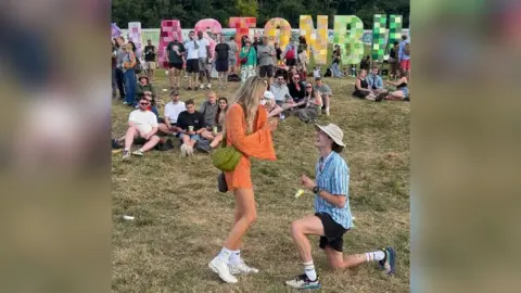 A man down on one knee as he proposes to his girlfriend in front of the Glastonbury sign.