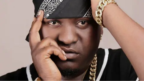 Close-up of musician Sefu Shabani in a black bandana with a white design posing with his hands near his face. He is wearing a chunky gold chain necklace and bracelet.