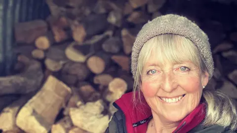 Kate MacRae A woman stands in front of a pile of wooden logs.