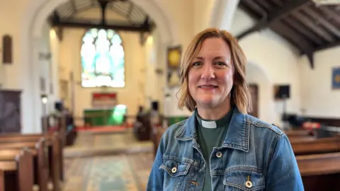 Reverend Elise Peterson inside St Mary and St Peter's Church in Wennington. The vicar has short blonde hair and is wearing a green top with a clerical collar and a denim jacket. In the background are rows of pews and a stained glass window.