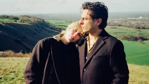 An image of a blonde woman who is wearing all black leaning on a man with brown hair who is also wearing all black in the middle of a field.