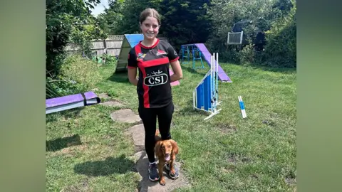 Kate Bradbrook/BBC Taylor with dark hair tied down smiling at the camera and wearing a red and black T-shirt and black trousers. A brown cocker spaniel is sitting at her feet. They are in a grassed garden where slides and ramps are arranged in a course.