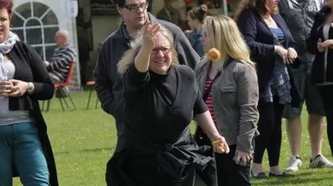 Woman in a field with several other people throwing a scone-like round biscuit. She is wearing glasses and has blonde hair.