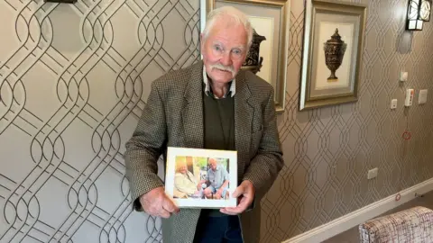 A man in a suit stands in a room and holds a photo in a frame. He has grey hair and a grey moustache. He is wearing a tweed jacket and a jumper. The framed photo shows him with a woman.