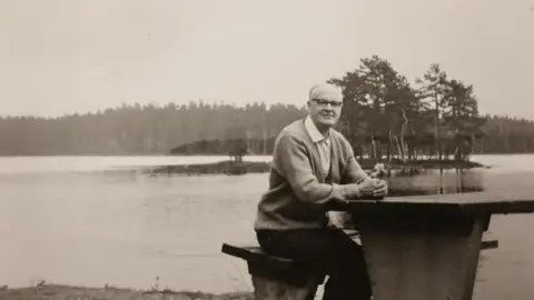 Michael Kitching A black and white photo of an older man witting on a bench. He has white hair and black glasses, and is wearing a white shirt, cardigan and dark trousers. Behind him is a lake with a small island of trees in the middle of it
