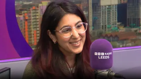 A person sits in a BBC Radio Leeds studio, speaking into a purple microphone with “BBC Radio Leeds” printed on it. The person has long dark hair and wears large, clear-framed glasses. Behind the person, a large window or backdrop shows a cityscape with modern buildings and a construction crane. Purple studio panels frame the scene.
