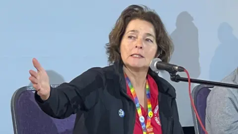 Local Democracy Reporting Service Caroline Hayhurst speaking into a microphone and lifting her arm as she sits on a purple chair on a panel. She has short brown hair and is wearing a rainbow lanyard with badges on it around her neck. She is also wearing a red polo shirt with a black jacket on top. 