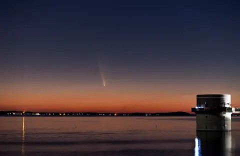 Reuters A building at the edge of a lake and then a settlement on the far side under an orange hue in the sky. Just above, in a bluer, higher portion of the sky, light from a comet can be seen.