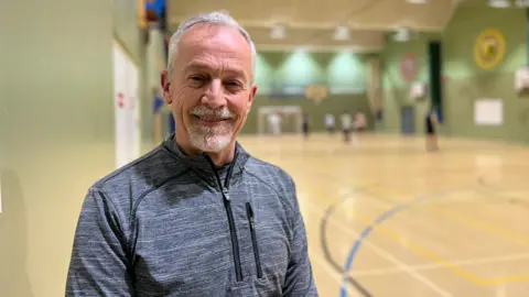 Gary smiles at the camera as the players are playing a game of futsal behind him. He is wearing a grey zip up fleece and has white, short hair and a white goatee.