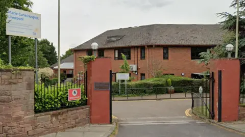 A sandstone brick entrance with the a white sign with blue writing which says Marie Curie Hospice, Liverpool. The entrance leads to a red brick building with dark framed windows surrounded by green bushes and trees.