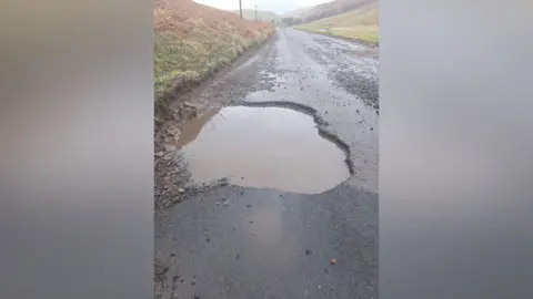 Malcolm Fairbairn A large and deep pothole on the side of a country road that extends to the middle of the carriageway. There is gravel strewn around the road with grassy hills on either side of the road. Hills can be seen in the distance.