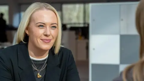 Sophie Branston, who is in a black blazer and who has shoulder-length blonde hair in conversation with another person, whose face is not visible, in an office setting.
