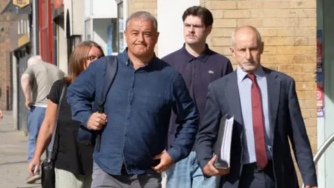 David Moore and other members of Emily's family walk, along with a bald bearded man in a suit carrying a large ring binder of documents, into the coroners court building in Crook. David Moore is clean shaven with short grey hair and a determined expression on his face. He is wearing a dark navy shirt. Behind him, partially seen, is a woman with shoulder length dark brown hair wearing glasses and a black short-sleeved top. Behind the suited man, who is on David's left, is a younger man with short dark hair in a centre parting. He has a moustache and wears a dark - perhaps purple - polo shirt and stonewashed blue jeans.