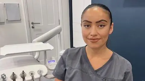 BBC Kath Stahl sits in her dental surgery. Her black hair is tied back and she wears grey scrubs. Dental equipment can be seen to her left. 