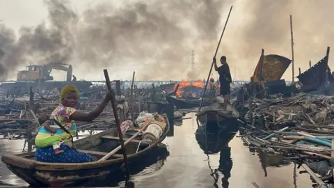 A woman with a baby on her back rows her boat through debris in Makoko. Another boat, a bulldozer and flames can be seen ahead of her.