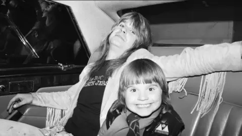 An old black and white photo of Ozzy and his son Louis. Ozzy is sat back with his arm stretched across small Louis who is smiling bright eyed and leaning forward towards the camera