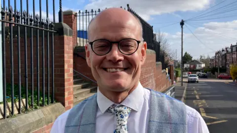 A bald man with round black glasses smiles at the camera. A road and a school fence extend down the street behind him. He has a flowery white tie, light lilac shirt and grey waistcoat.