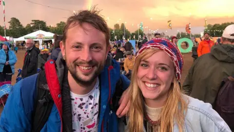 Jody White A picture of Jody White and Jessie White. Jody is wearing a blue and red jacket. He is carrying a a black bag. Jessie is wearing a light blue denim jacket and a crochet head band. They are both at a festival, 