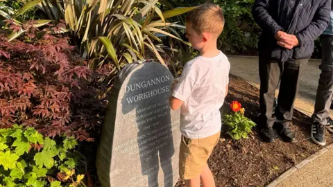 A young boy stands at the memorial stone which is in a garden space surrounded by various plants and bushes. The young boy has short light hair and is looking at the stone with his hands clasped, wearing a white t-shirt and light shorts.