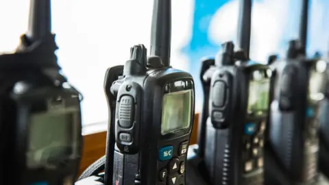 A row of four black walkie talkies/radios are all placed upwards against a window with one in focused.