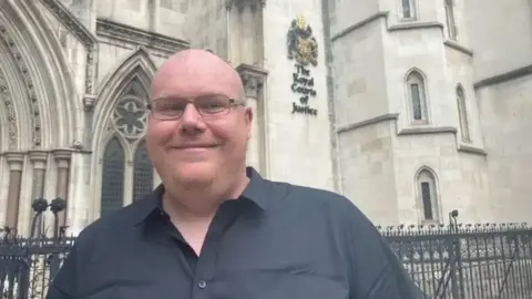 Mr Brentnall wearing glasses and a black shirt standing outside the Royal Courts of Justice. There is a black wrought iron fence behind him and an ornate arched window. 