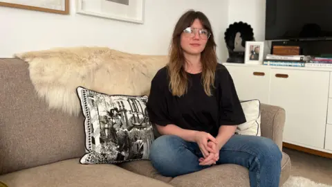 Abi looks at the camera as she sits on a grey sofa as she wears blue jeans and a black t-shirt. She has dark brown hair with lighter ends which comes just past her shoulders. She is sitting next to a cushion with black and white print pillow.