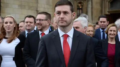 PA Jim McMahon, que tem cabelo curto e escuro e barba por fazer, usa terno e gravata vermelha. Ele está no centro do quadro com um grupo de políticos e apoiadores trabalhistas, incluindo Angela Rayner, na foto atrás dele.