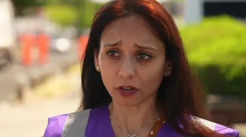 A woman in a purple high-visibility vest speaks outdoors on a sunny street.