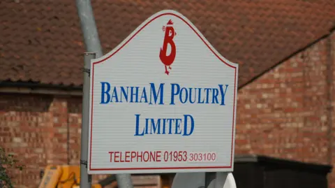 We can see the Banham Poultry Sign. There's a red B logo at the top - with chicken features - the company name beneath it in blue - and its phone number below.