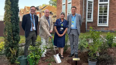 Four people in a garden planting a tree with a brick building behind them.