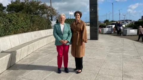 A woman with white hair and a patterned green blazer with red trousers, stands next to a woman with brown hair in a brown coat, in front of a grey monument. 