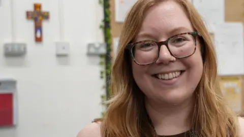 Sarah Horspole standing in a primary school classroom. She is wearing a brown sleeveless top and is looking directly at the camera and smiling.
