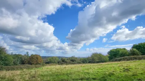 A sunny day looking out over a field lined with trees. The sky is blue but filled with some white clouds. Hills can be seen in the distance