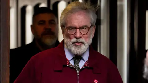 A man with grey hair and white beard, he is wearing brown glasses, a red jacket, blue shirt and green tie with white dots. He has a red circle pin with a white circle on it on his chest