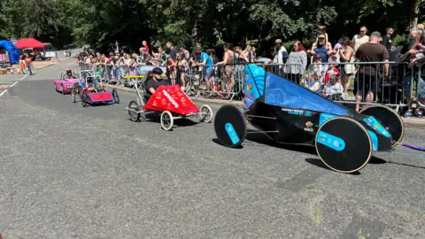 Four colourful karts racing in a line on a road. Crowds can be seen behind the metal barriers. 