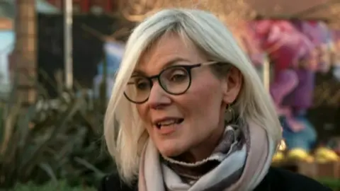 Nora Smith who has short blonde hair and is standing outside in a cream and pink scarf and a black coat/jacket. She has black rimmed glasses on. 