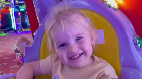 PA Media Little girl smiling at the camera, sitting in a brightly coloured chair at an amusement arcade