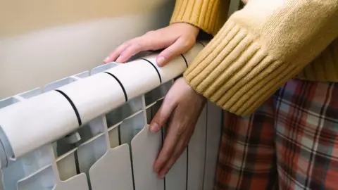 Getty Images A stock photo of the arms and legs of a person wearing pyjamas and a yellow jumper warming their hands on a radiator. 