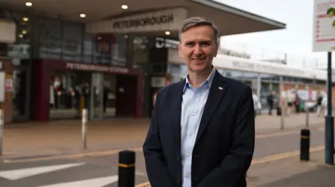 Andrew Pakes stares at the camera. He wears a black suit and a light blue shirt. He stands in front of a road with double yellow lines and speed hump. Behind that is a building which says Peterborough station.