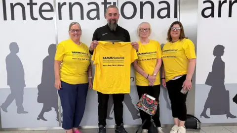 Eugene Scardifield Three smiling women and a man wearing bright yellow charity T-shirts standing in front of an airport arrivals sign
