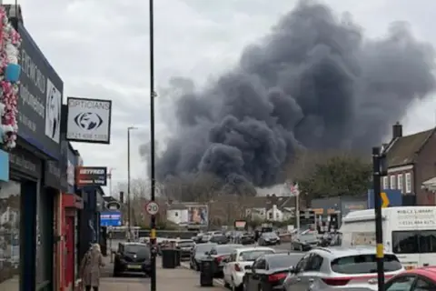 WMFS Grey smoke seen in the sky of a residential street lined with cars. There is a lamppost with a 20 mph sign on it as well as a green large rubbish bin. An elderly woman has her back to the camera in the distance.