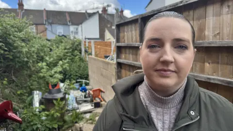 Gabby Colenso/BBC Laura Haydon, wearing a green anorak over a grey ribbed sweater, looks directly at the camera. Her hair is swept back and she has blue eyes. Behind her are a fence and some dumped rubbish, including a child's scooter and a plastic chair, and the backs of some terraced houses.