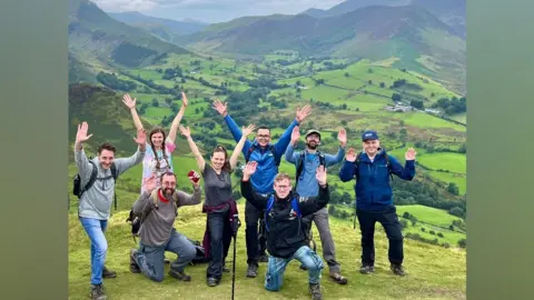 Hannah Morley The Stag Walkers posing for a photo on a hill, with mountains in the background. There are nine people with their hands raised in the air, smiling. Two kneel at the front. There are some trees in the valley and a few scattered houses.