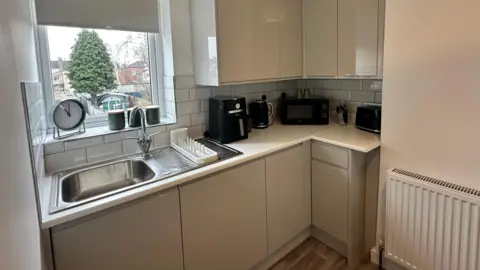 A modern-looking kitchenette, with a window overlooking a garden dominated by an evergreen tree. The kitchen cupboards are cream-coloured and there is an aluminium basin. A black microwave, kettle and other kitchen gadgets stand on the counter.