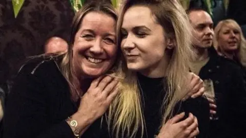 Mary Curran (left) holds her daughter Lily Lucas by her shoulders, hugging and smiling together. They are both wearing black outfits and Lily has long blonde hair and her mother has brown hair and is waring a bracelet. A man and woman can be seen in the background. 