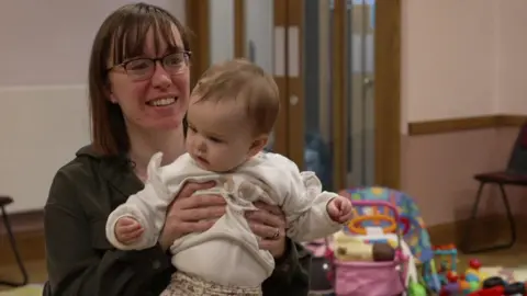 A mother smiling whilst holding her baby up at a baby class.
