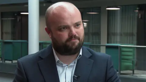 Close up image of Llŷr Powell standing in the BBC building in Cardiff. He is wearing a light blue shirt and navy blazer which has a small microphone attached on the left collar. He is looking at the interviewer not the camera. Powell is bald with a brown beard and has a stern expression. 