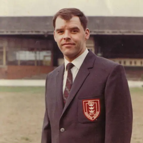 Hull KR Graham Paul wearing a Hull Kingston Rovers club blazer and tie he has short dark hair and is standing on the pitch in front of a stand